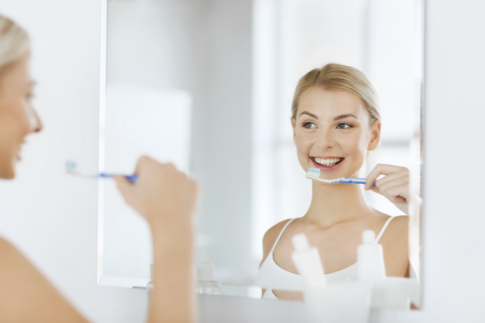 health care, dental hygiene, people and beauty concept - smiling young woman with toothbrush cleaning teeth and looking to mirror at home bathroom