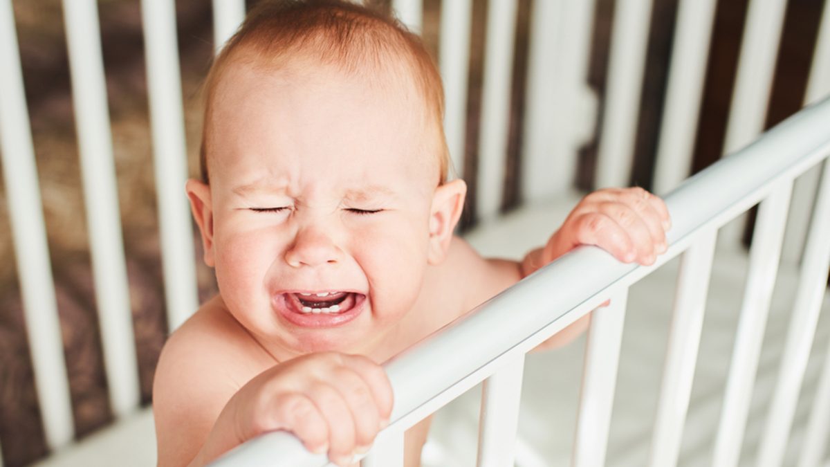 A year-old child is crying in the crib and holds onto the side of the bed